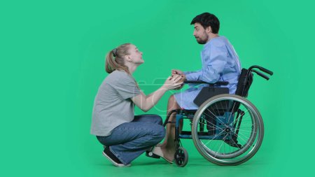 Photo for Medical ward and rehabilitation creative concept on chroma key green screen. Adult man patient in robe sitting in wheelchair girl next to him on knees holding his hand, supporting expression. - Royalty Free Image