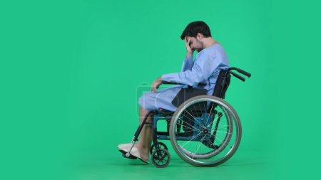 Photo for Medical ward and rehabilitation creative concept on chroma key green screen. Adult man patient in robe sitting in wheelchair holding head in hand, looking at his legs, depressed expression sad face. - Royalty Free Image