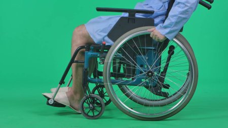 Photo for Medical ward and rehabilitation creative concept on chroma key green screen. Adult man patient in robe sitting in wheelchair and riding on it helping to spin the wheels with hands, close up shot. - Royalty Free Image