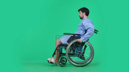 Photo for Medical ward and rehabilitation creative concept on chroma key green screen. Adult man patient in robe sitting in wheelchair and riding on it helping to spin the wheels with hands. - Royalty Free Image