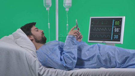 Photo for Medical ward and rehabilitation creative concept on chroma key green screen. Adult man patient laying in bed with drip and monitor, holding smartphone, watching social media online. - Royalty Free Image