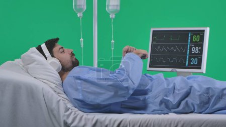 Photo for Medical ward and rehabilitation creative concept on chroma key green screen. Adult man patient laying in bed with drip and monitor, wearing headphones listening music on smartphone. - Royalty Free Image