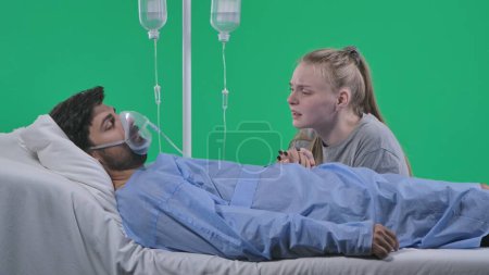 Photo for Medical ward and rehabilitation creative concept on chroma key green screen. Adult man patient laying in bed with drip and mask, female sitting next to man holding hand, crying face expression. - Royalty Free Image
