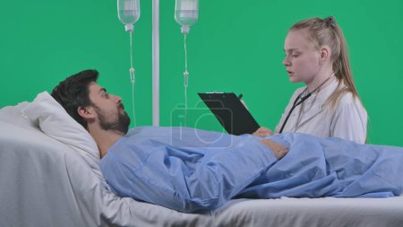 Photo for Medical ward and rehabilitation creative concept on chroma key green screen. Adult man patient laying in bed with drip, female nurse sitting next to man with clipboard asking questions talking. - Royalty Free Image