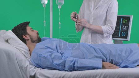 Photo for Medical ward and rehabilitation creative concept on chroma key green screen. Adult man patient laying in bed with drip and monitor, female nurse draws injection in syringe holding in hand. - Royalty Free Image