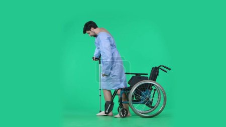 Téléchargez les photos : Service médical et concept créatif de réhabilitation sur écran vert chromatique. Homme adulte patient en robe debout du fauteuil roulant, faisant quelques pas tenant la béquille à la main pour soutenir le corps. - en image libre de droit