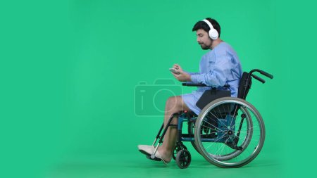 Photo for Medical ward and rehabilitation creative concept on chroma key green screen. Adult man patient in robe sitting in wheelchair wearing headphones holding smartphone, listening to music. - Royalty Free Image