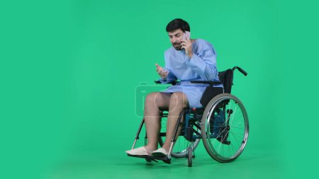 Photo for Medical ward and rehabilitation creative concept on chroma key green screen. Adult man patient in robe sitting in wheelchair holding smartphone and talking, positive face expression and gesture. - Royalty Free Image