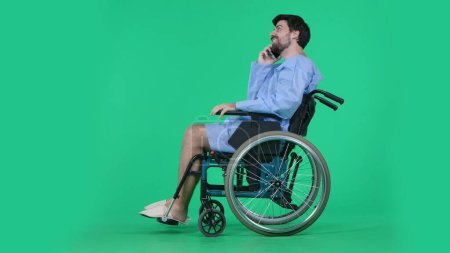 Photo for Medical ward and rehabilitation creative concept on chroma key green screen. Adult man patient in robe sitting in wheelchair holding smartphone and talking, positive smiling face expression. - Royalty Free Image