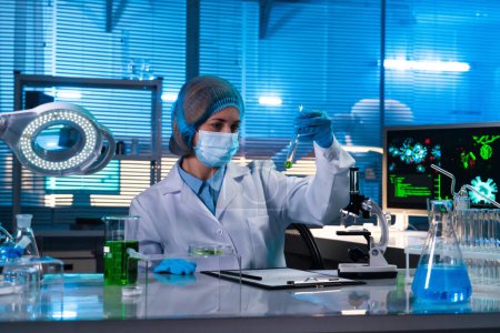 Foto de Científica sentada en un laboratorio de investigación. Una doctora con una bata blanca estudiando una muestra de líquido verde en un tubo de ensayo. El concepto de un laboratorio de bioquímica - Imagen libre de derechos