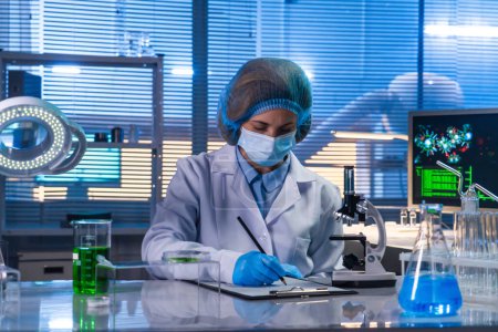 Photo for Female scientist sitting in a research lab. A female doctor in a white coat writing down research data on a sheet. The concept of a bio or chemical laboratory - Royalty Free Image