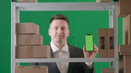 Photo for Framed on a green background, chromakey. Depicts an adult male in a suit. Demonstrates a manager, a supervisor in a warehouse. He holds a phone with a green screen to the camera and smiles happily. - Royalty Free Image
