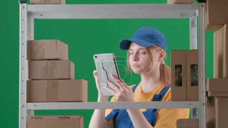 Photo for Framed on a green background, chromakey. Depicts a young woman in uniform and cap. Demonstrates an employee, a storekeeper in a warehouse. She holds a clipboard and scrutinizes, examines boxes. - Royalty Free Image