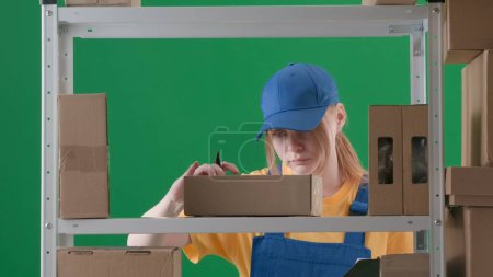 Photo for Framed on a green background, chromakey. Depicts a young woman in uniform and cap. Demonstrates an employee, a storekeeper in a warehouse. She is attentive, serious and signs boxes with a marker. - Royalty Free Image