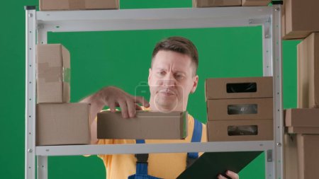 Photo for Framed on green background, chromakey. Depicted is an adult male wearing a work uniform. Demonstrates a storekeeper in a warehouse. He holds a clipboard with his other hand a box and looks carefully. - Royalty Free Image