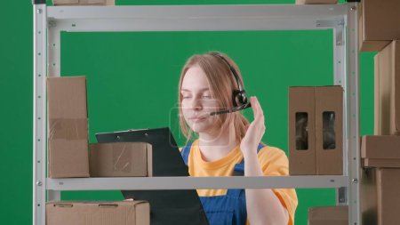 Photo for Framed on a green background, chromakey. Depicts a young woman wearing a uniform and headphones. Demonstrates a worker, a storekeeper in a warehouse. She holds a tablet and looks at something. - Royalty Free Image