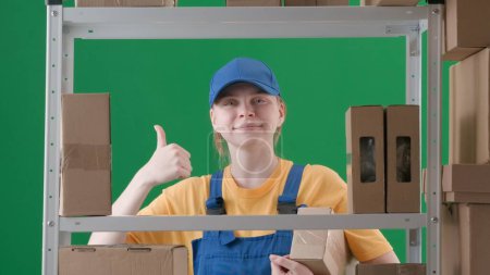 Photo for Framed on a green background, chromakey. Depicts a young woman wearing a uniform and cap. Demonstrates a worker, a storekeeper in a warehouse. She holds in a box, smiles and shows a gesture Cool. - Royalty Free Image