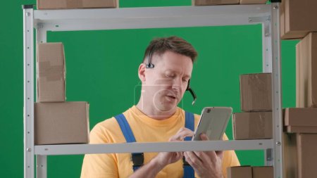 Photo for Framed on green background, chromakey. Depicted is an adult male wearing a work uniform. Demonstrates a storekeeper in a warehouse. He is wearing headphones and looking intently at a tablet. Medium. - Royalty Free Image