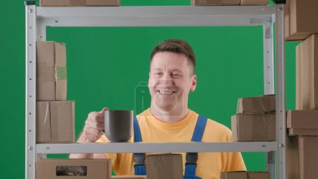 Téléchargez les photos : Encadré sur fond vert, chromatique. Un homme adulte portant un uniforme de travail est représenté. Démontre un magasinier dans un entrepôt. Il tient une tasse avec un verre et sourit, il est heureux, satisfait - en image libre de droit