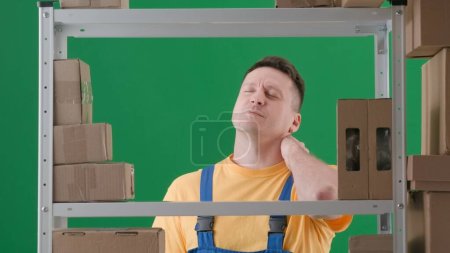 Photo for Framed on green chromakey background. Adult man in work uniform. Depicts a storekeeper in a warehouse. He unhappily kneads his neck and arms, he is tired of work, wants to rest. - Royalty Free Image