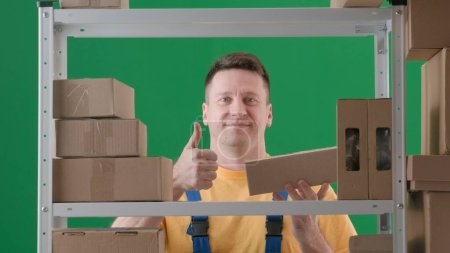 Photo for In the frame on a green background, chromakey. Adult man in work uniform. Demonstrates the storekeeper in a warehouse. He is got a box in his hand, and the other one is doing a cool gesture and - Royalty Free Image