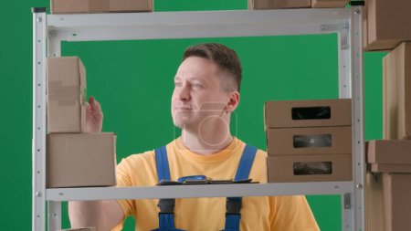 Photo for Framed on green background, chromakey. Depicted is an adult male wearing a work uniform. Demonstrates a worker, a storekeeper in a warehouse. He scrutinizes the boxes and smiles. Medium frame. - Royalty Free Image