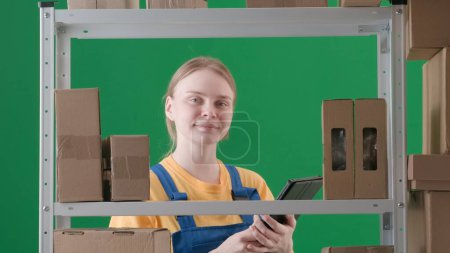 Photo for Framed on a green background, chromakey. Depicts a young woman in uniform. Demonstrates a worker, a storekeeper in a warehouse. She looks into the camera, smiles happily and holds a clipboard. - Royalty Free Image