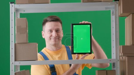 Téléchargez les photos : Encadré sur fond vert, chromatique. Un homme adulte portant un uniforme de travail est représenté. Démontre un magasinier dans un entrepôt. Il tient une tablette avec l'écran face à la caméra et souriant - en image libre de droit