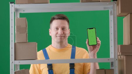 Téléchargez les photos : Encadré sur fond vert, chromatique. Un homme adulte portant un uniforme de travail est représenté. Démontre un magasinier dans un entrepôt. Il tient son téléphone avec l'écran face à la caméra et sourit. - en image libre de droit