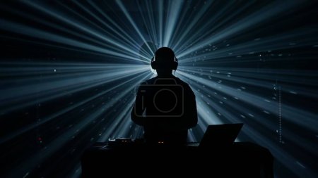 Foto de Una silueta de un DJ masculino con auriculares se levanta contra un telón de fondo iluminado de vibrantes luces de escenario en un evento del club. La atmósfera es eléctrica, con haces de luz azul y verde que atraviesan - Imagen libre de derechos