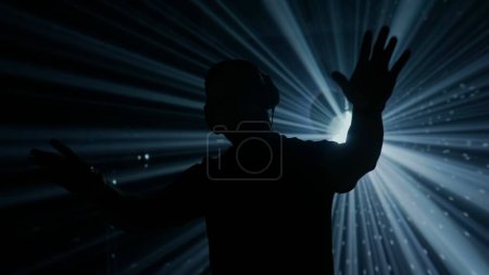 Téléchargez les photos : Une silhouette d'un DJ masculin avec écouteurs se dresse sur une toile de fond éclairée de lumières de scène vibrantes lors d'un événement du club. L'atmosphère est électrique, avec des rayons de lumière bleue et verte coupant à travers - en image libre de droit
