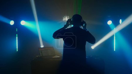 Foto de Una silueta de un DJ masculino con auriculares se levanta contra un telón de fondo iluminado de vibrantes luces de escenario en un evento del club. La atmósfera es eléctrica, con haces de luz azul y verde que atraviesan - Imagen libre de derechos