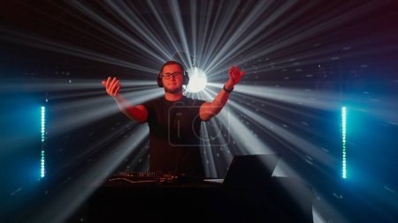 Photo for Captivating professional DJ at work in a nightclub setting, with hands on deck controlling sound mixer and turntables. The focused male DJ, wearing headphones, expertly mixes tracks during a live set - Royalty Free Image