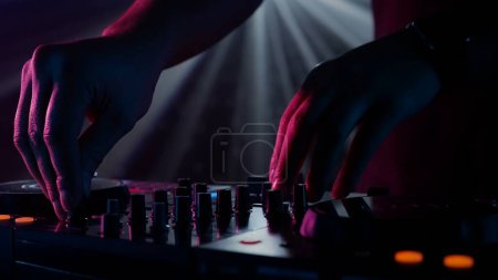 Téléchargez les photos : La photo zoome sur les mains habiles d'un DJ ajustant les boutons d'un mixeur sonore, avec une toile de fond d'éclairage du club ambiant. Le contraste vibrant des mains rouges contre les tons frais de vert et - en image libre de droit