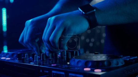 Téléchargez les photos : La photo zoome sur les mains habiles d'un DJ ajustant les boutons d'un mixeur sonore, avec une toile de fond d'éclairage du club ambiant. Le contraste vibrant des mains rouges contre les tons frais de vert et - en image libre de droit