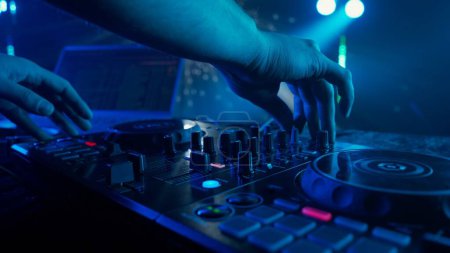 Photo for The photo zooms in on the skilled hands of a DJ adjusting the knobs of a sound mixer, with a backdrop of ambient club lighting. The vibrant contrast of red hands against the cool tones of green and - Royalty Free Image