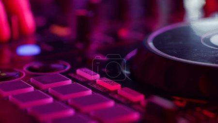 Foto de Esta imagen de primer plano muestra un tablero de mezclas de DJ bañado en luces del club de neón, destacando los intrincados detalles del equipo de sonido. Rosas vívidas y púrpuras proyectan un resplandor en el tocadiscos y botones - Imagen libre de derechos