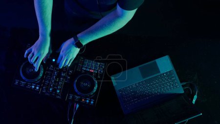 Das Foto zoomt auf die geschickten Hände eines DJs, der die Regler eines Soundmischpults vor dem Hintergrund der Ambient-Club-Beleuchtung justiert. Der lebhafte Kontrast roter Hände zu den kühlen Tönen von Grün und Grün