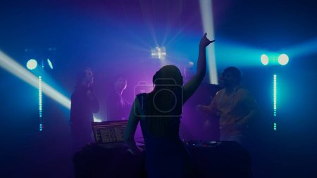 Foto de La imagen captura a un DJ con un brazo levantado, liderando la carga en una vibrante discoteca. La multitud se ve en silueta, inmersa en la experiencia, con rayos de luz radiantes en el fondo - Imagen libre de derechos