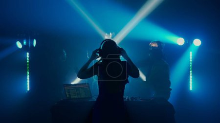 Foto de La imagen captura a un DJ con un brazo levantado, liderando la carga en una vibrante discoteca. La multitud se ve en silueta, inmersa en la experiencia, con rayos de luz radiantes en el fondo - Imagen libre de derechos
