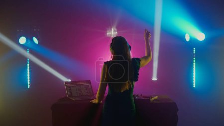 Téléchargez les photos : L'image capture le moment exubérant d'une DJ féminine levant la main dans la joie, rétro-éclairée par un spectre de lumières de scène colorées dans une boîte de nuit. Shes portant des écouteurs et face à la caméra - en image libre de droit