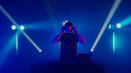 Téléchargez les photos : DJ professionnel captivant au travail dans un cadre de boîte de nuit, avec les mains sur le pont contrôlant mélangeur sonore et platines tournantes. Le DJ masculin concentré, coiffé d'écouteurs, mixe habilement les morceaux lors d'un live set - en image libre de droit
