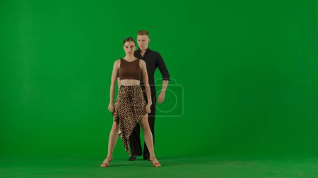 Foto de Un elegante dúo de baile de salón realiza una rutina dinámica capturada sobre un vívido fondo de pantalla verde. El bailarín masculino, vestido con un elegante traje negro, golpea una postura equilibrada con una mano - Imagen libre de derechos