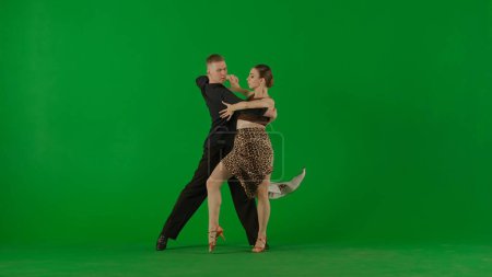 Téléchargez les photos : Un élégant duo de danse de salon exécute une routine dynamique capturée sur un fond vert vif. Le danseur masculin, vêtu d'une élégante tenue noire, frappe d'une main - en image libre de droit