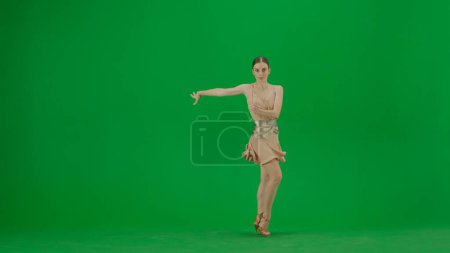 Foto de Bailarina de salón latina en Pose Against Green Screen. Una bailarina de salón latina equilibrada se levanta contra un telón de fondo de pantalla verde, su forma exudando elegancia y control. Vestido con un vestido de baile beige con - Imagen libre de derechos