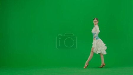 Photo for Latin Ballroom Dancer in Pose Against Green Screen. A poised Latin ballroom dancer stands against a green screen backdrop, her form exuding elegance and control. Clad in a beige dance dress with - Royalty Free Image
