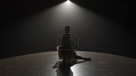 Téléchargez les photos : La silhouette saisissante d'une danseuse est capturée sous un seul projecteur sur une scène sombre, transmettant un moment d'intensité dramatique. La lumière projette une longue ombre, ajoutant de la profondeur à la scène et - en image libre de droit