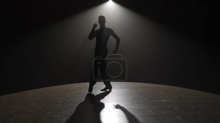 Photo for Silhouette Elegant Ballroom Dancer on Spotlight. The striking silhouette of a male dancer is captured under a single spotlight on a dark stage, conveying a moment of dramatic intensity. The light - Royalty Free Image