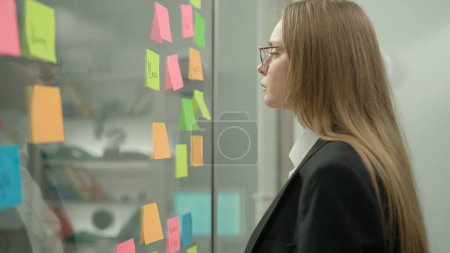 Photo for Brainstorming and ideas in modern business creative concept. Portrait of corporate leader in office planning project on notes. Business woman next to glass wall full of sticky notes. - Royalty Free Image