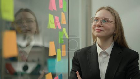 Photo for Brainstorming and ideas in modern business creative concept. Portrait of corporate leader in office planning project on notes. Business woman next to glass wall full of sticky notes, smiling face. - Royalty Free Image